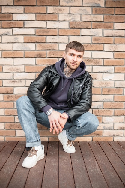 Cool trendy hipster guy in fashion casual clothes with leather jacket jeans hoodie and sneakers sits near a brick wall on the street