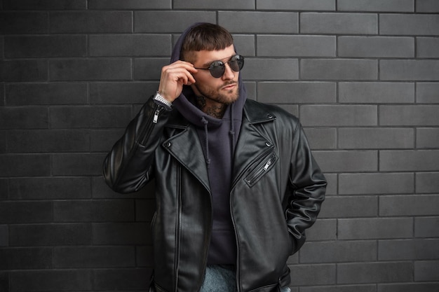 가죽 재킷과 후드티를 입은 세련된 옷을 입은 멋진 힙스터 모델 남자는 검은 벽돌 벽 근처에서 세련된 선글라스를 조정합니다