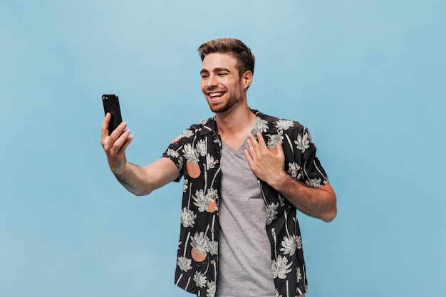 Cool trendy man in good mood with stylish beard in grey plaid t-shirt and summer dark shirt taking selfie and smiling on blue wall