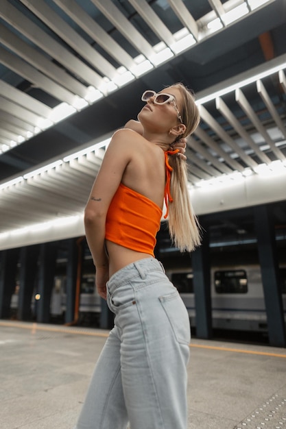 Cool urban fashion damesmodel met coole zonnebril in trendy vrijetijdskleding met oranje top en vintage jeans in de metro