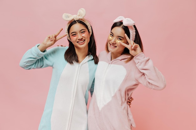 Cool young Asian women in pajamas show peace signs on pink background Girls in kigurumis and headbands pose with cosmetic eyepatches