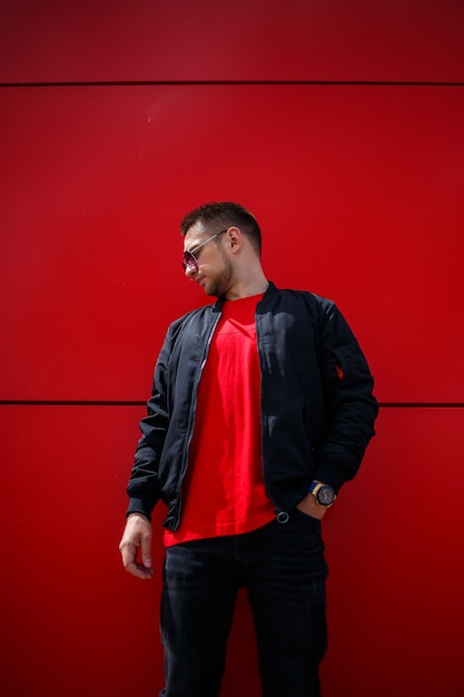 A cool young man in trendy sunglasses in a stylish red T-shirt with a black jacket stands near a red building in the city. Attractive modern guy. Fashionable summer men's clothing. Street style