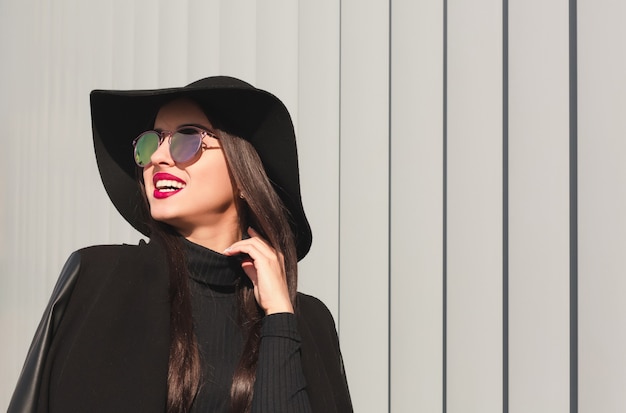 Cool young model  in mirror glasses and broad brimmed hat posing near the city building