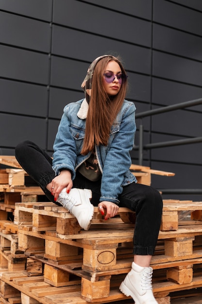 Cool young stylish hipster woman in trendy casual youth clothes in fashionable purple glasses poses in the city. American sexy glamorous girl fashion model sits on wooden pallets in the street. Style.