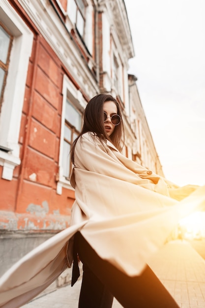 Cool young woman in fashion sunglasses in fashionable spring beige trench coat posing on street near building at sunset. Pretty lovely urban girl model enjoys walk and orange bright sunlight outdoors.