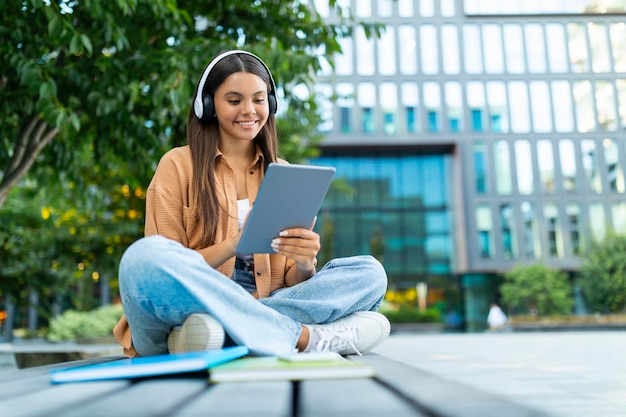 Cool young woman student sitting outdoors using digital tablet headphones