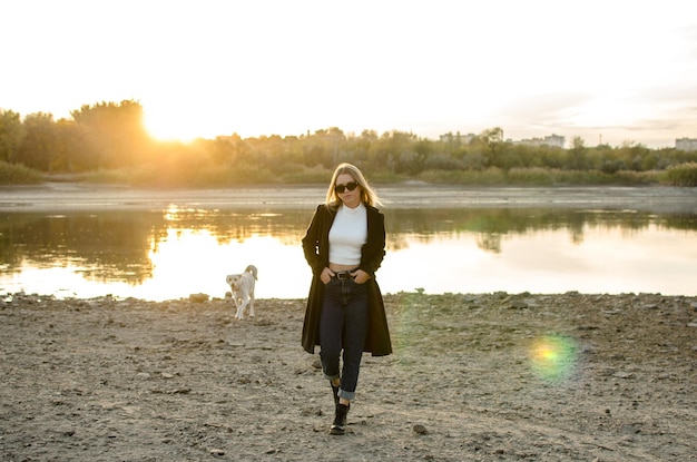 A cool young woman in sunglasses and a black coat walks her dog on the riverbank at sunset