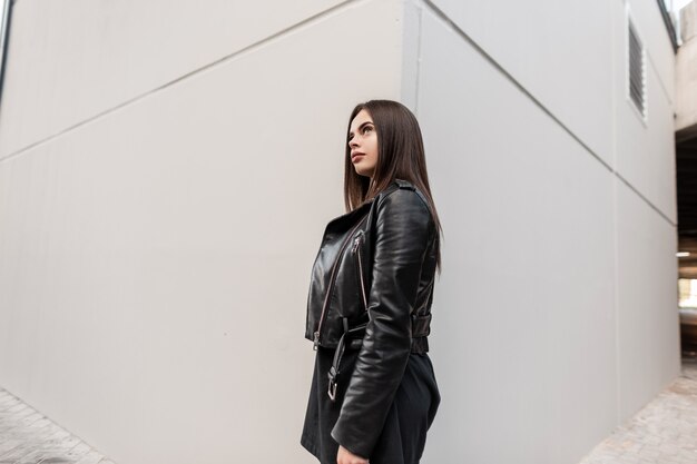 Coole jonge, mooie stijlvolle hipster-vrouw in een zwart leren jasje en jurk in de buurt van een grijze moderne muur op straat
