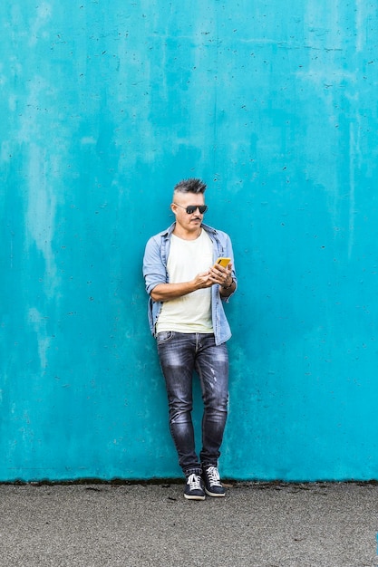 Coole man kijkt naar mobiele telefoon die buiten tegen een blauwe muur leunt Man verslaafd aan technologie en sociale netwerken Verticaal en kopieer ruimte voor tekst