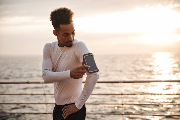 Coole man met donkere huidskleur in wit t-shirt met lange mouwen en zwarte korte broek tikt op het telefoonscherm en traint in de buurt van zee