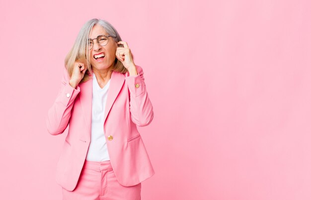 Coole vrouw van middelbare leeftijd die boos, gestrest en geïrriteerd kijkt en beide oren bedekt met een oorverdovend geluid, geluid of luide muziek tegen een vlakke muur