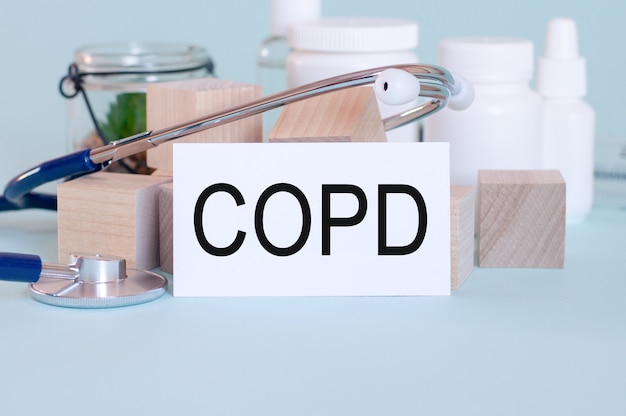 Слова Copd, написанные на белой медицинской карте, со стетоскопом, зеленым цветком, медицинскими таблетками и деревянными блоками на синем фоне