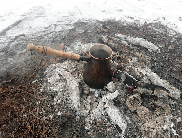 A copper cezve with coffee is cooked on the hot coals of a fire\
and there is snow around it