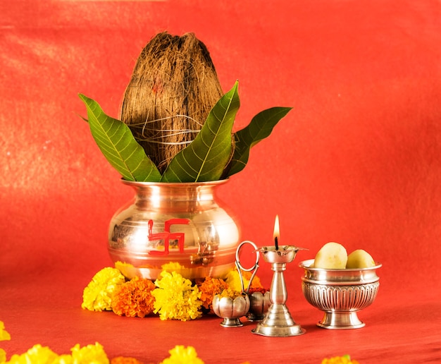 Photo copper kalash with coconut and mango leaf with floral decoration, diya, haldi kumkum and sweet pedha,essential in hindu puja, front view, closeup on red surface