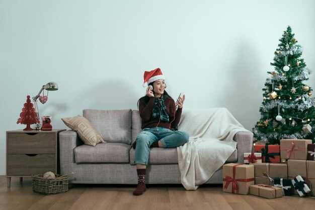 복사 공간입니다. 크리스마스 시간에 집 거실에서 헤드폰을 끼고 온라인 휴대전화로 음악을 들으며 평온한 아시아 일본 여성. 산타 모자를 쓰고 따뜻한 실내에서 스웨터를 입은 소녀는 노래를 즐깁니다.