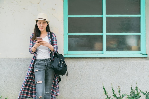 copy space concept. beautiful young asian chinese woman standing and leaning on white wall relax texting message on cellphone. lady with bag waiting for someone by window using mobile phone online