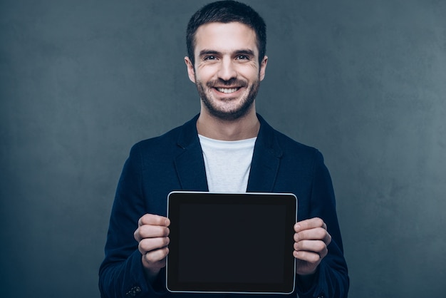 彼のデジタルタブレットのスペースをコピーします。彼のデジタルタブレットを示し、灰色の背景に立って笑っている陽気な若い男