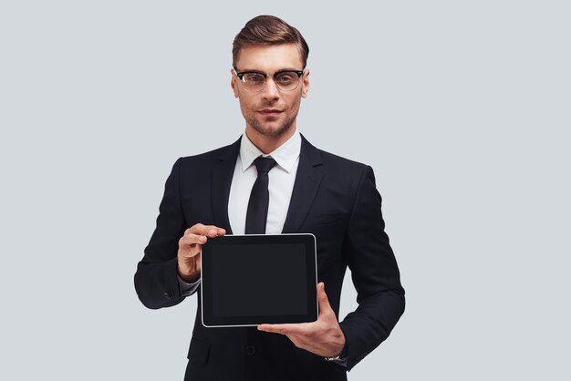 Copy space on his digital tablet. Good looking young man in full suit holding digital tablet and looking at camera while standing against grey background