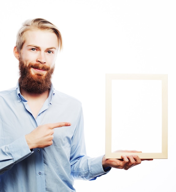 Copy space in picture frame. Handsome young bearded man in blue shirt holding a picture frame and pointing it with smile while standing isolated on white background