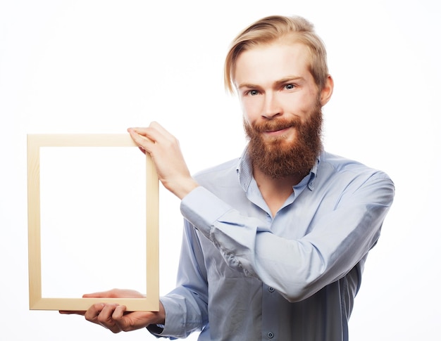 Copy space in picture frame Handsome young bearded man in blue shirt holding a picture frame and pointing it with smile while standing isolated on white background
