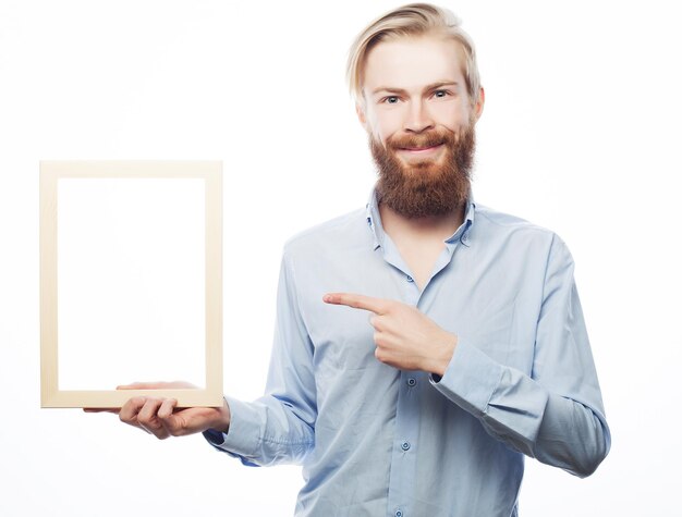 Copy space in picture frame Handsome young bearded man in blue shirt holding a picture frame and pointing it with smile while standing isolated on white background