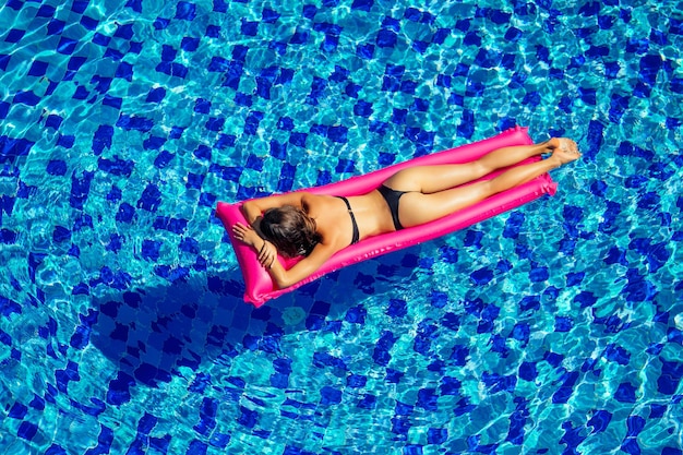 Copia spazio spf e crema solare bella ragazza bruna che galleggia nell'acqua della piscina. donna che nuota e si rilassa sul materasso gonfiabile rosa in piscina blu lavoro a distanza e vista dall'alto freelance