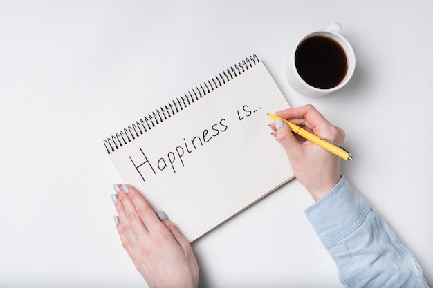 Copybook with HAPPINESS IS text. Top view of female hands writing in Notebook, cup of coffee