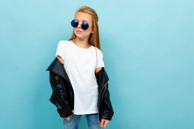 Copyspace-foto van cool stijlvol schoolmeisje op een blauwe achtergrond in een wit t-shirt en een zwarte jas