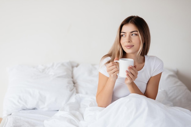 Copyspace-foto van een rustige blonde vrouw die opzij kijkt terwijl ze in haar bed zit en een koffiekopje vasthoudt