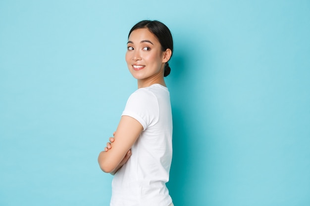 Coquettish pretty asian girl in white t-shirt standing in profile and turning right with curious smiling face, looking at something interesting, found good promo offer, standing blue wall