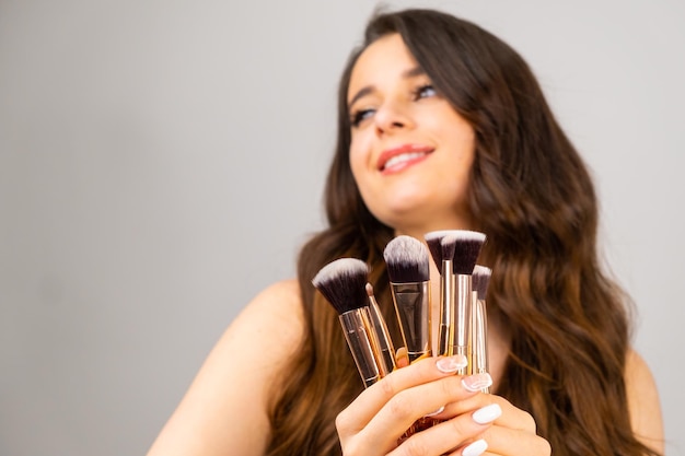 Coquettish young girl holding cosmetic brushes in her hands on the grey background with copy space