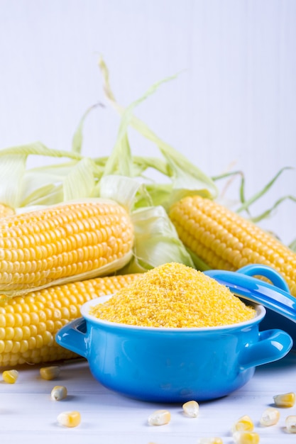 Photo corn grits polenta in a blue ceramic bowl on white table with ripe raw corn cob and green leaves