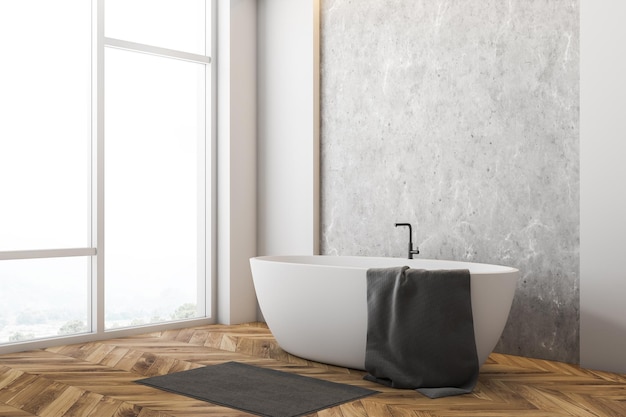 Corner of minimalistic bathroom with white and concrete walls, wooden floor, large window and white bathtub with towel on it and gray rug near it. 3d rendering