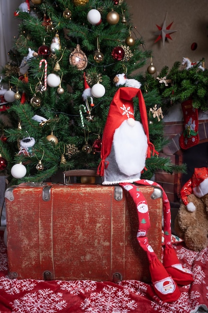 На углу красного старого чемодана сидит гном возле украшенной рождественской елки