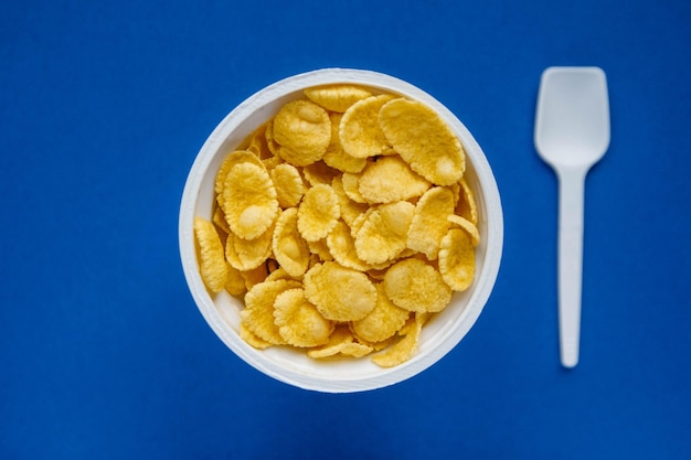Cornflakes in white bowl with spoon on blue background. Top view. Copy, empty space for text