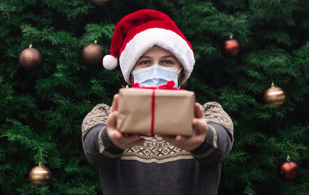 コロナクリスマスおめでとうございます。医療マスクでサンタの帽子とセーターを着て、赤いリボン、背景にクリスマスツリーのボケ味のギフトプレゼントボックスを与える肖像画の男