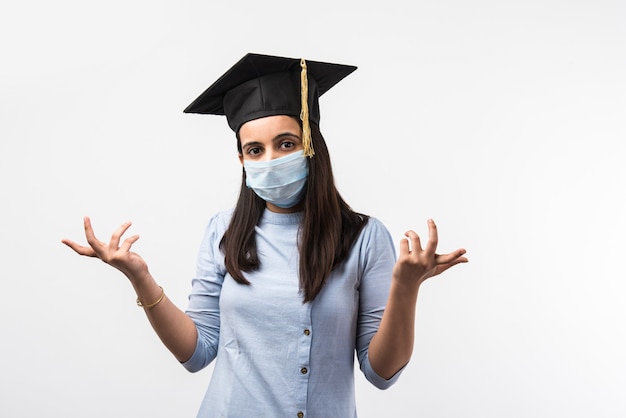 Corona Pandemic and confusion over university exams in India - Pretty Indian Female student with confused expressions wearing medical face mask and graduation hat