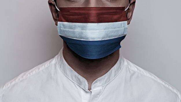 코로나 바이러스 세계적 대유행. 코로나 바이러스 검역, 코비드-19의 개념. 남성의 얼굴은 네덜란드 국기 색으로 칠해진 보호용 의료 마스크로 덮여 있습니다