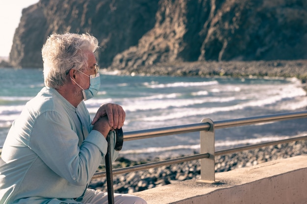 Photo coronavirus. bearded old man sitting on the beach in the sunset light holding a walking stick because of body aches