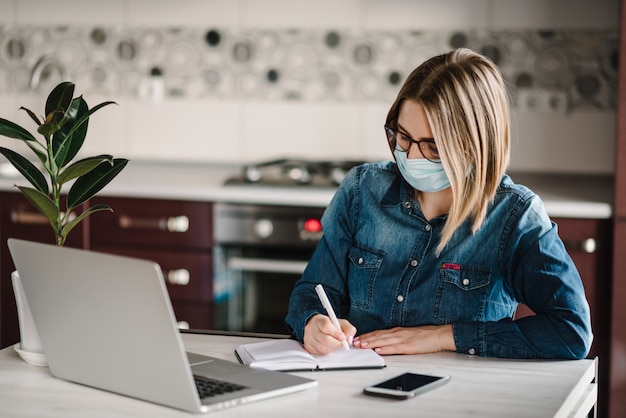 Coronavirus. Bedrijfsvrouw die in huis werken, die beschermend masker in quarantaine dragen. Blijf thuis. Het meisje leert, gebruikend laptop computer in huisbureau. Freelance. Schrijven, typen.