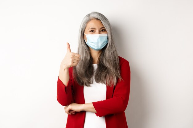 Coronavirus and business concept. Asian female entrepreneur in face mask looking cheerful, showing thumb-up in approval, praising using protective measures at work.