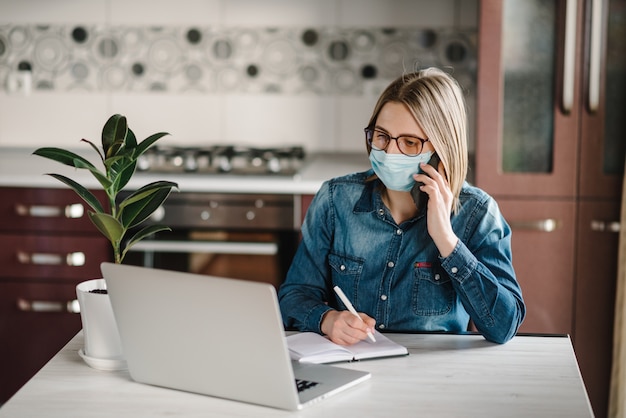 Coronavirus. Business woman working, talking on phone, wearing protective mask in quarantine. Stay at home. Girl learns, using laptop computer in home office. Freelance. Writing, typing.