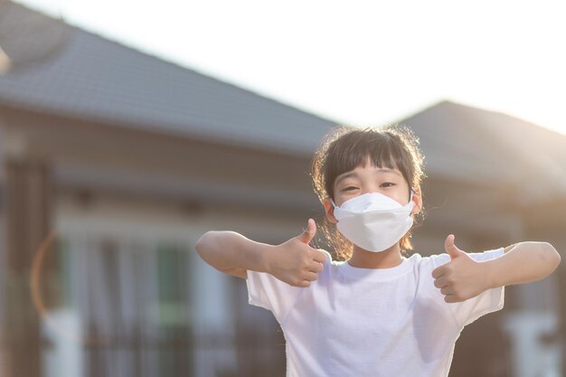 コロナウイルスコビッド-19pm2.5。オンライン教育。フェイスマスクを身に着けている小さな中国の女の子は、家で良いと幸せのために親指を表示します。
