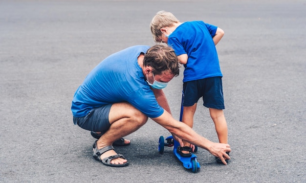 Coronavirus COVID 19 Sport active outdoor family life Father son wearing surgical sterilizing face mask dad explains technique of setting foot when riding scooter asphalt workout together