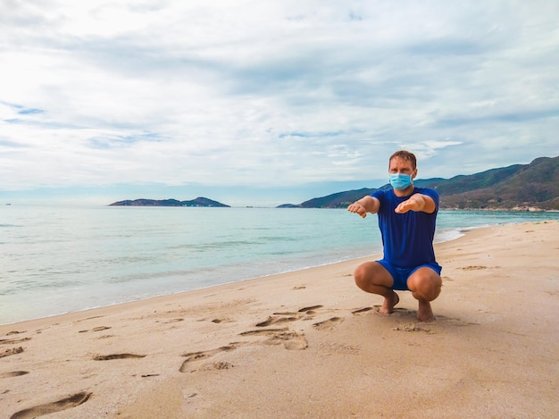コロナウイルスCOVID19検疫中の朝、トレーニングエクササイズを実行し、海の近くでしゃがむスポーツをしている若い美しい男サージカル滅菌フェイスマスク保護のアクティブな生活