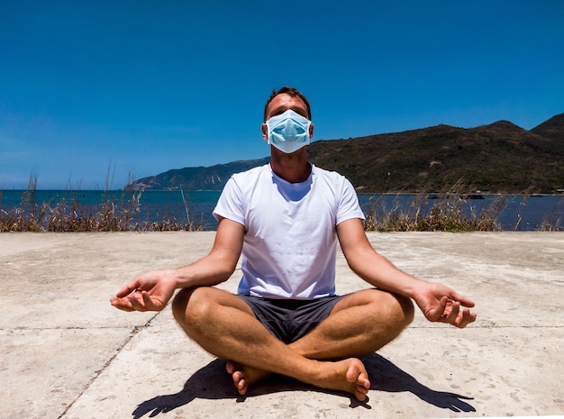 Coronavirus covid 19 young beautiful man doing sport performing\
workouts exercises yoga in the park near sea during quarantine\
active life in surgical sterilizing face mask protection