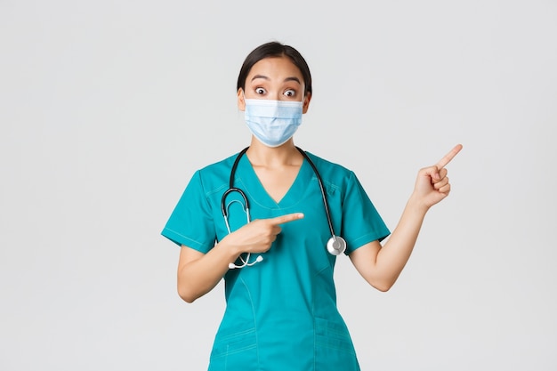 , coronavirus disease, healthcare workers concept. Enthusiastic happy asian female doctor, physician or nurse in medical mask and scrubs, pointing upper right corner, show awesome promo.