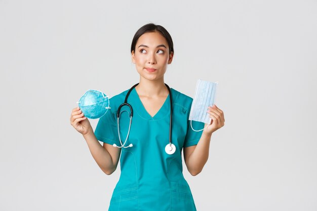 , coronavirus disease, healthcare workers concept. Thoughtful smiling asian female doctor, physician in scrubs looking dreamy upper left corner, holding respirator and medical mask.