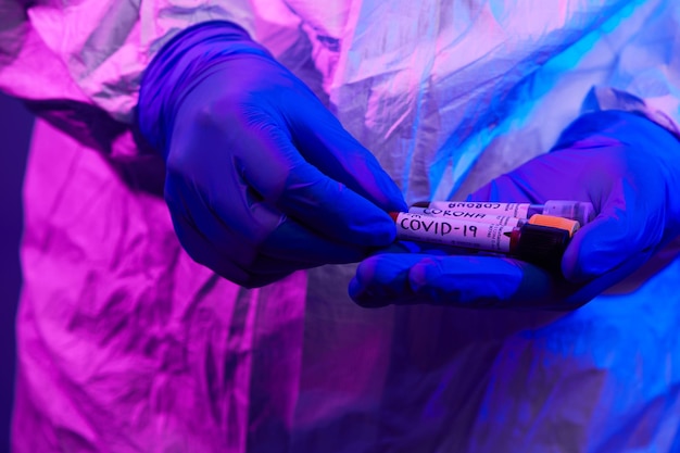 Coronavirus, doctor holding positive covid 19 virus blood sample test tube. wearing biohazard epidemic protective mask, suit and glows neon light background