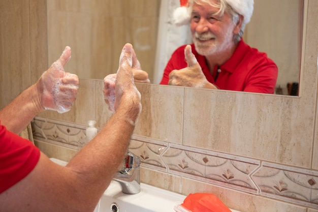 Coronavirus. een gelukkige senior man met een kerstmuts wast zijn handen thuis met een ok teken met zijn hand. rood chirurgisch masker om coronavirusinfectie op de gootsteen te voorkomen